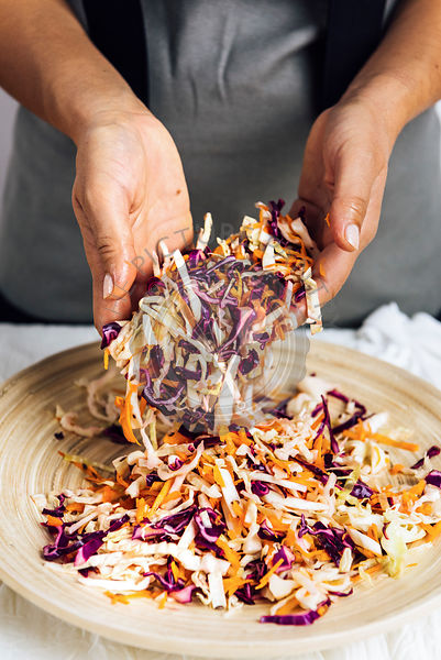 A woman tossing shredded carrot, white and purple cabbage in a wooden bowl photographed from front view.